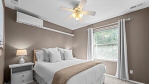 A bedroom with a bed, bedside table, lamp, ceiling fan, window, and curtains.