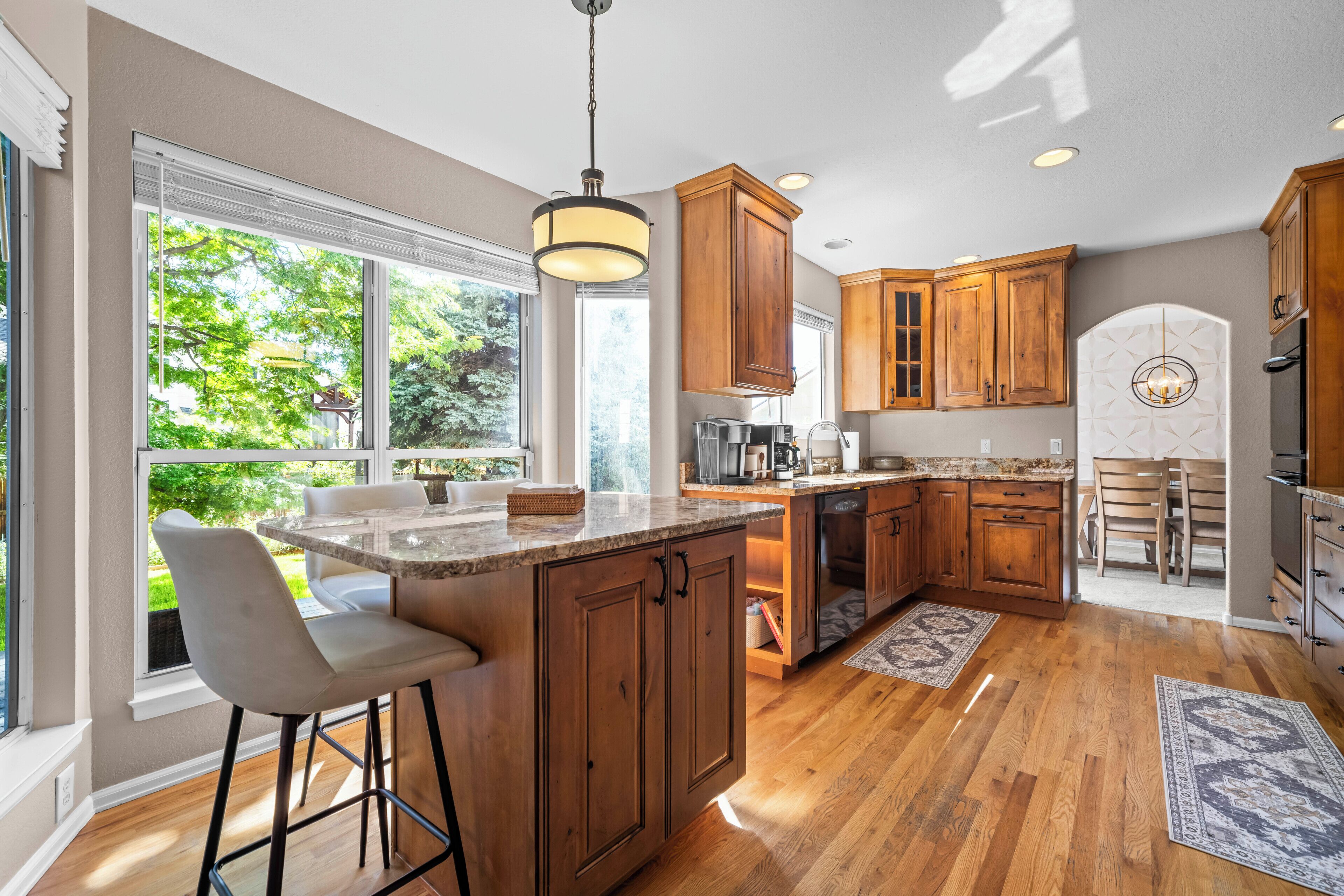 A kitchen with wooden cabinets, a marble countertop, and a large window overlooking greenery.