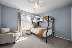 A bedroom with a bunk bed, a bedside table, a chair, and a window with curtains.