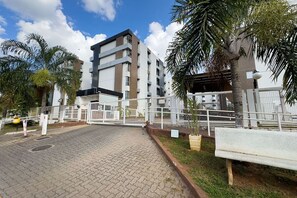 Una entrada porticada a un edificio moderno, con un camino pavimentado, palmeras y un banco.