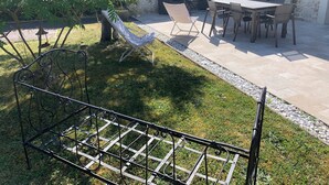 Un patio con mesa y sillas, una parrilla metálica y un jardín con un árbol y una hamaca.