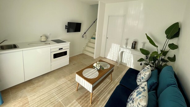 Cocina moderna con horno empotrado, un lavamanos y una tetera. Hay una zona de comedor con mesa y sillas, y un sofá con un cojín estampado. Un televisor está montado en la pared, y se ve una escalera al fondo.