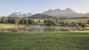 Um lago com um píer, rodeado por campos gramados e árvores, com montanhas ao fundo.