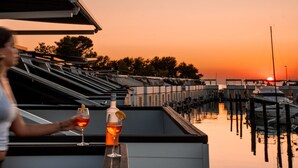 Henkilö, jolla on kädessään lasi punaviiniä ja toisessa kädessään pullo, seisoo katolla, jolta avautuu näkymä venesatamaan ja auringonlaskuun.