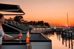 Una persona sosteniendo una copa de vino tinto y una botella en una azotea con vista a un puerto deportivo y al atardecer.