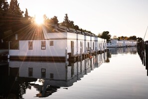 Una fila di case mobili bianche con finestre, collegate a un pontile sull'acqua, con il sole che splende sullo sfondo.