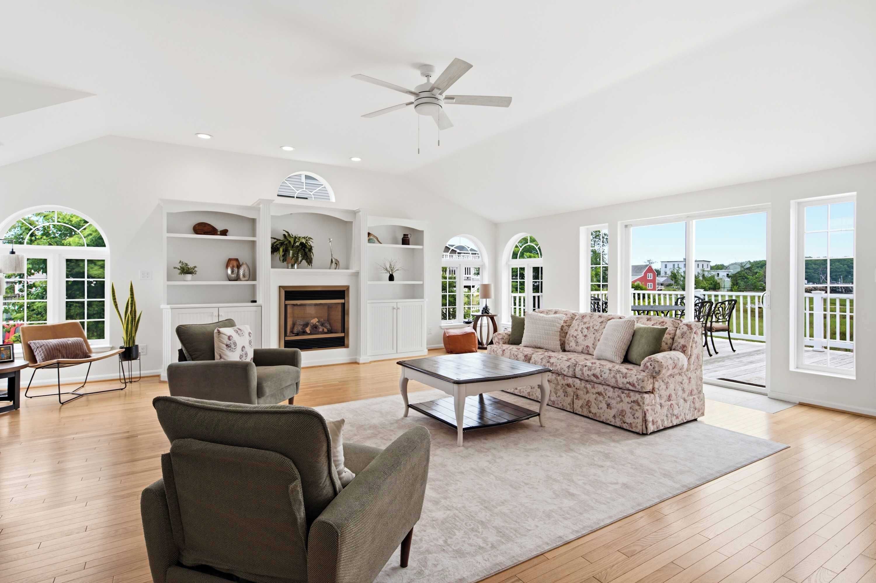 A spacious living room with a fireplace, a ceiling fan, and large windows offering a view of the outdoors.