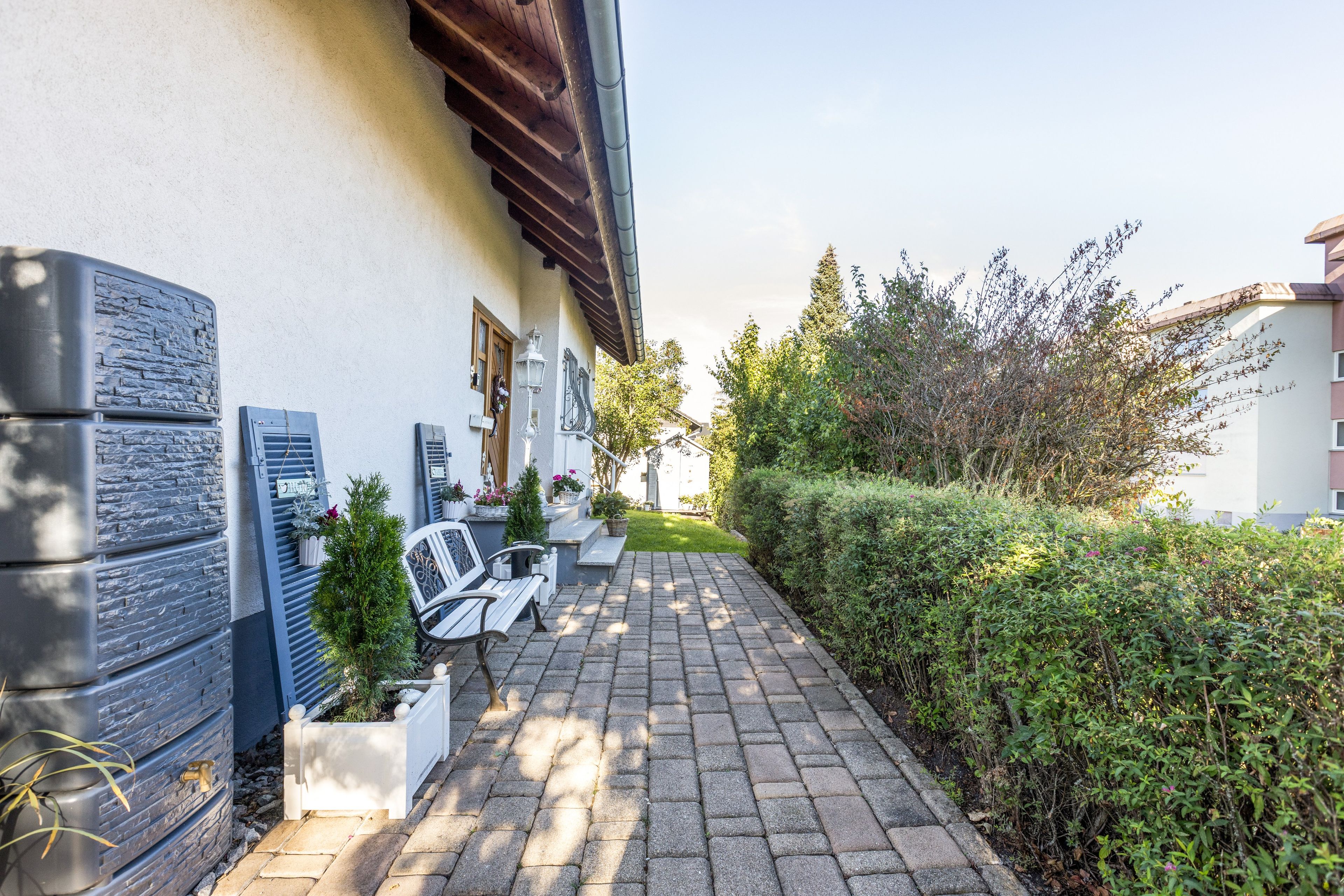 Ein gepflasterter Weg führt zu einem Gebäude mit weißer Fassade, einer Holztür und einem kleinen Gartenbereich mit Blumentöpfen und einer Bank.