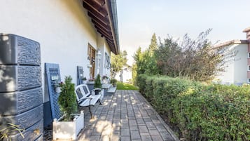 Ein gepflasterter Weg führt zu einem Gebäude mit weißer Fassade, einer Holztür und einem kleinen Gartenbereich mit Blumentöpfen und einer Bank.