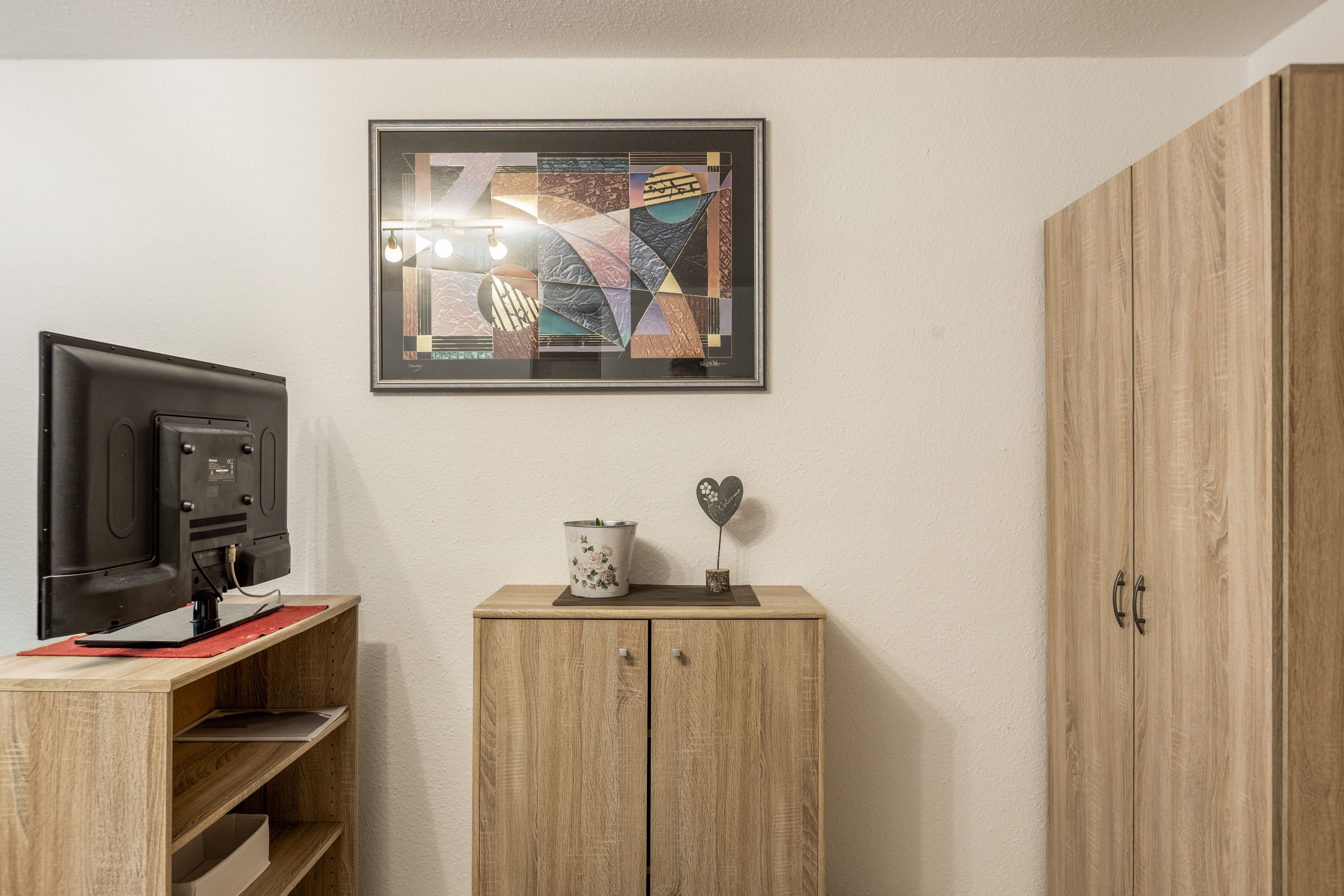 Quarto com televisão em um suporte, um armário com um vaso de flores e uma obra de arte abstrata emoldurada na parede.