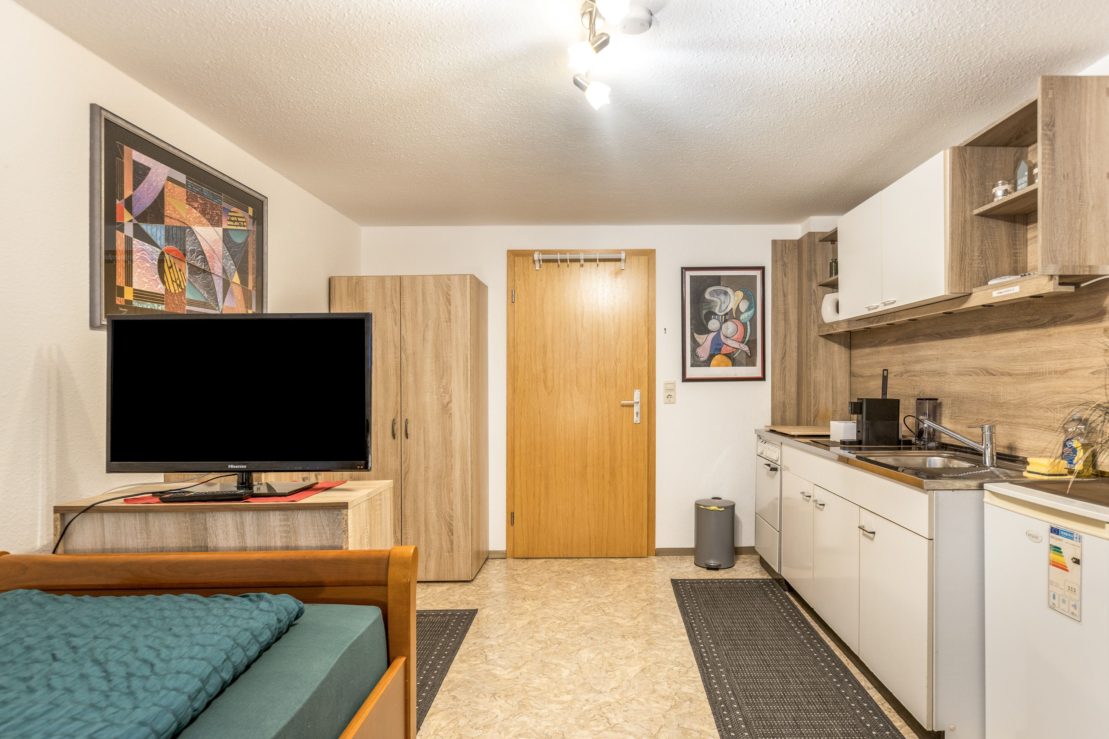 Een kleine hotelkamer met een bed, een televisie, een houten garderobe, een kitchenette en een deur naar een andere kamer.