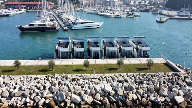 Una marina con diverse imbarcazioni ormeggiate, un molo roccioso e una fila di edifici moderni con tetti piani.