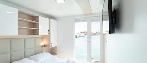 Una habitación de hotel con cama, televisor montado en la pared, ventana con vista al mar y estante empotrado.