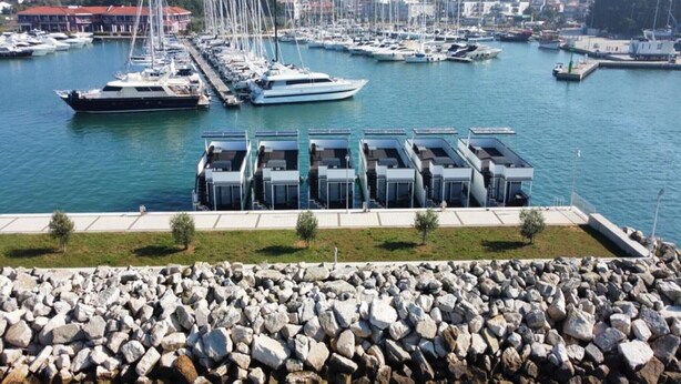 Uma marina com vários barcos atracados, um paredão rochoso e uma fileira de edifícios modernos com telhados planos.