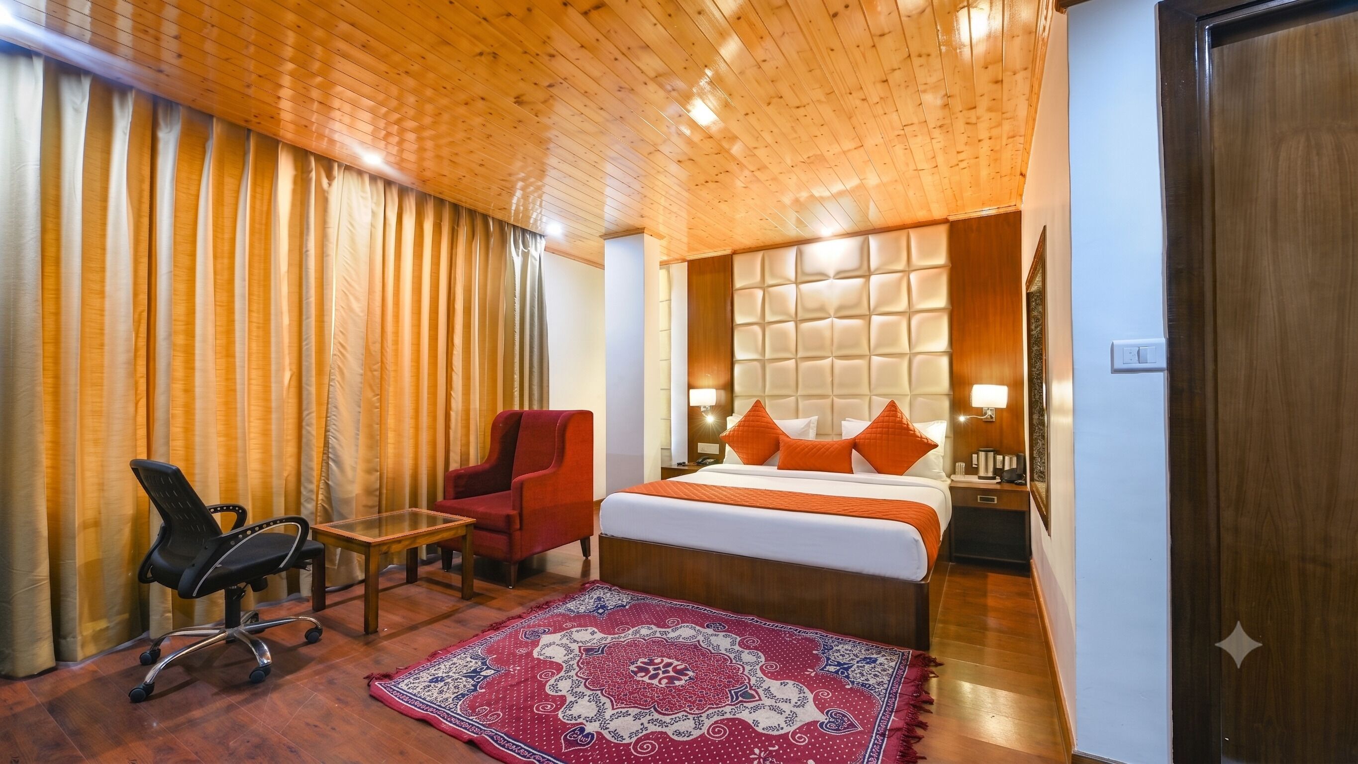Une chambre d’hôtel avec un plafond en bois, un lit avec une literie orange, un fauteuil rouge, une chaise de bureau noire, une petite table et un tapis à motifs.