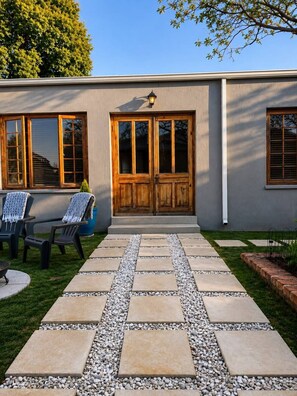 A patio with a stone pathway leading to a double wooden door, outdoor furniture, and a small tree.