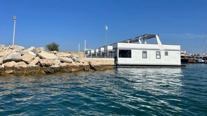 Uma moderna casa flutuante atracada em uma marina, com costa rochosa e águas azul-claras.