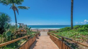 A pathway leading to a beach with a wooden fence, a sign, and palm trees. - Ap en Cond. c/ Piscina a 6 min de la Playa Gsh0102 (Camaçari)