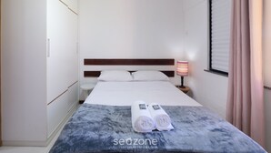 A hotel room with a bed, a bedside table with a lamp, a window with curtains, and a towel on the bed. - Ap en Cond. c/ Piscina a 6 min de la Playa Gsh0102 (Camaçari)