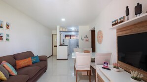 A modern living room with a brown sofa, white dining table, and a flat-screen TV. - Ap en Cond. c/ Piscina a 6 min de la Playa Gsh0102 (Camaçari)