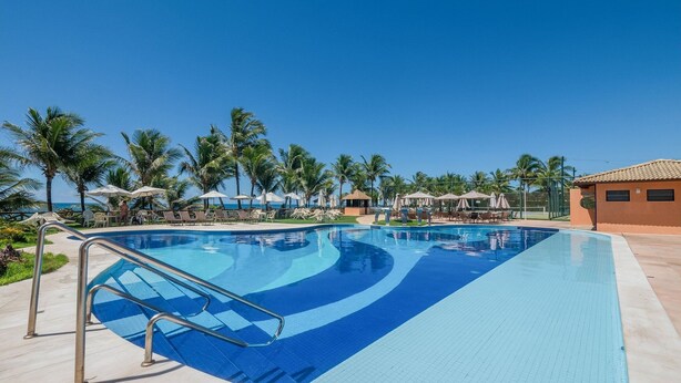 A swimming pool with a blue and white tiled design, surrounded by lounge chairs and umbrellas, with palm trees and a clear sky in the background. - Apt in a Seaside Condominium With a Pool - Gsh2101 (Camaçari)