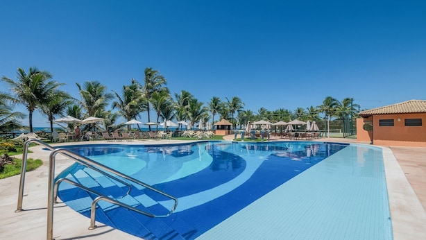 A swimming pool with a blue and white tiled design, surrounded by lounge chairs and umbrellas, with palm trees and a clear sky in the background. - Apt in Condo With Pool Near the Beach Gsh0101 (Camaçari)