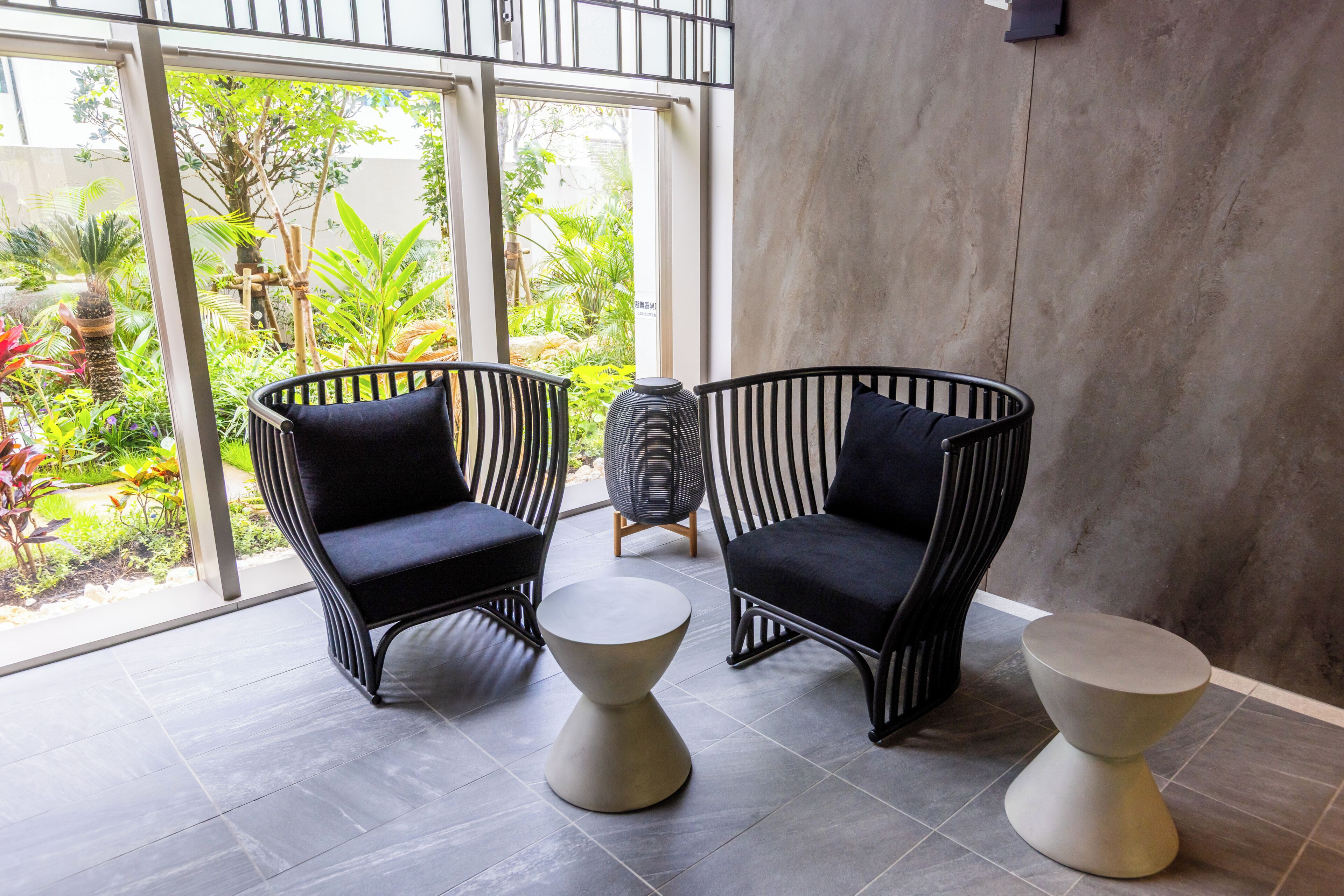 A modern indoor space with two black wicker chairs with cushions, a small round table, and a large window offering a view of a garden.
