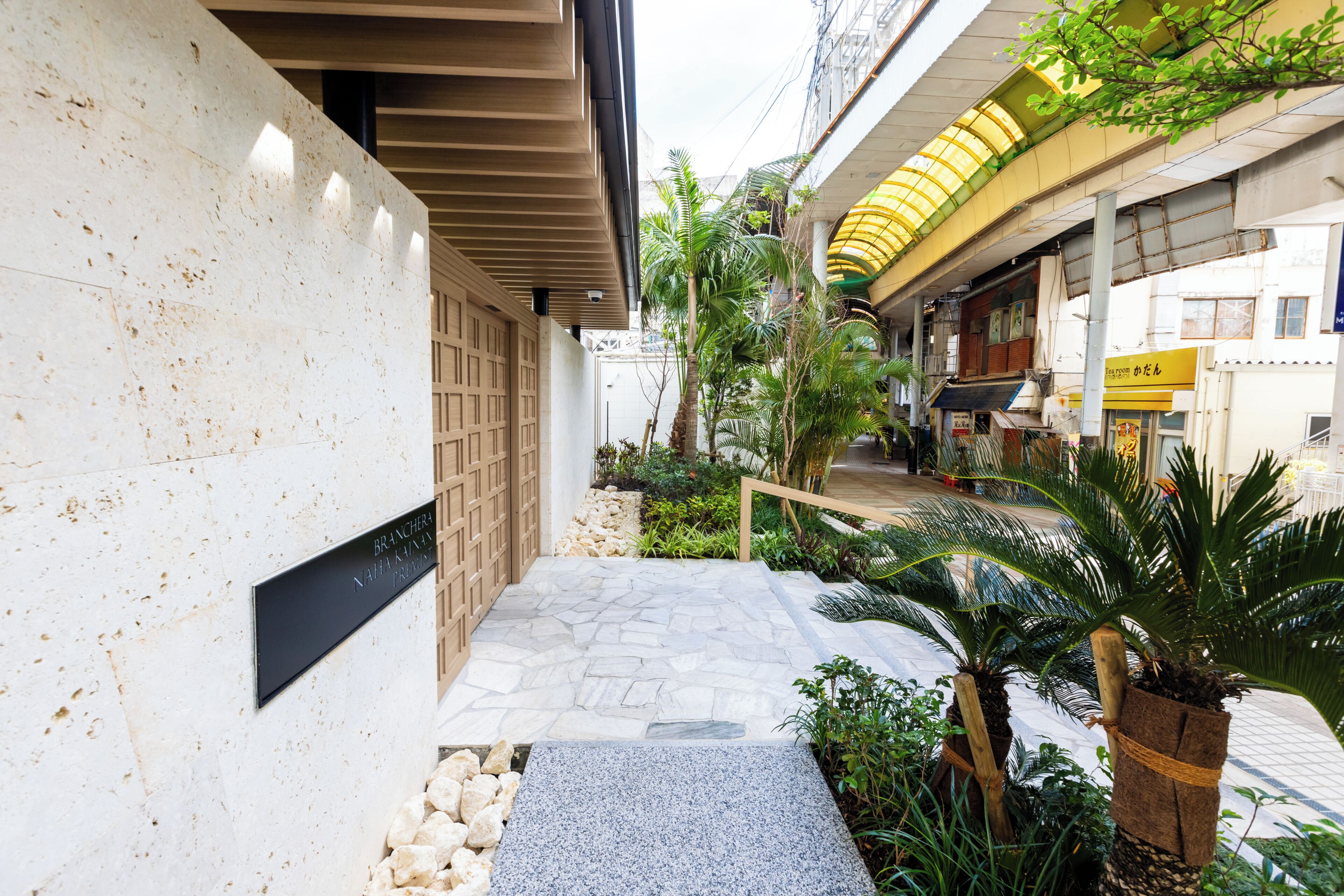 A narrow pathway with stone flooring leading to a building entrance, flanked by greenery and a wooden fence.