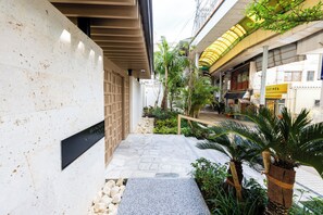 Un étroit sentier dallé de pierres menant à l’entrée d’un bâtiment, bordé de verdure et d’une clôture en bois.