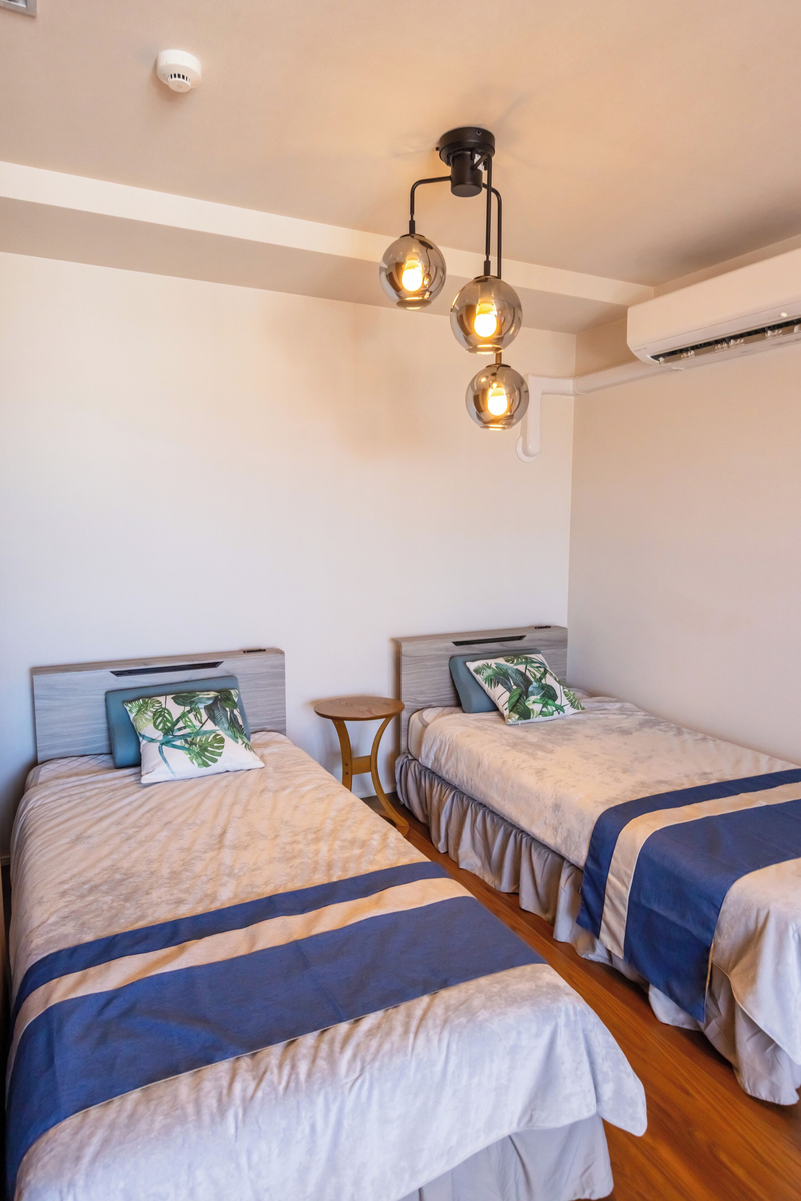 A room with two beds, each with a headboard, a small round table, and a ceiling light fixture.