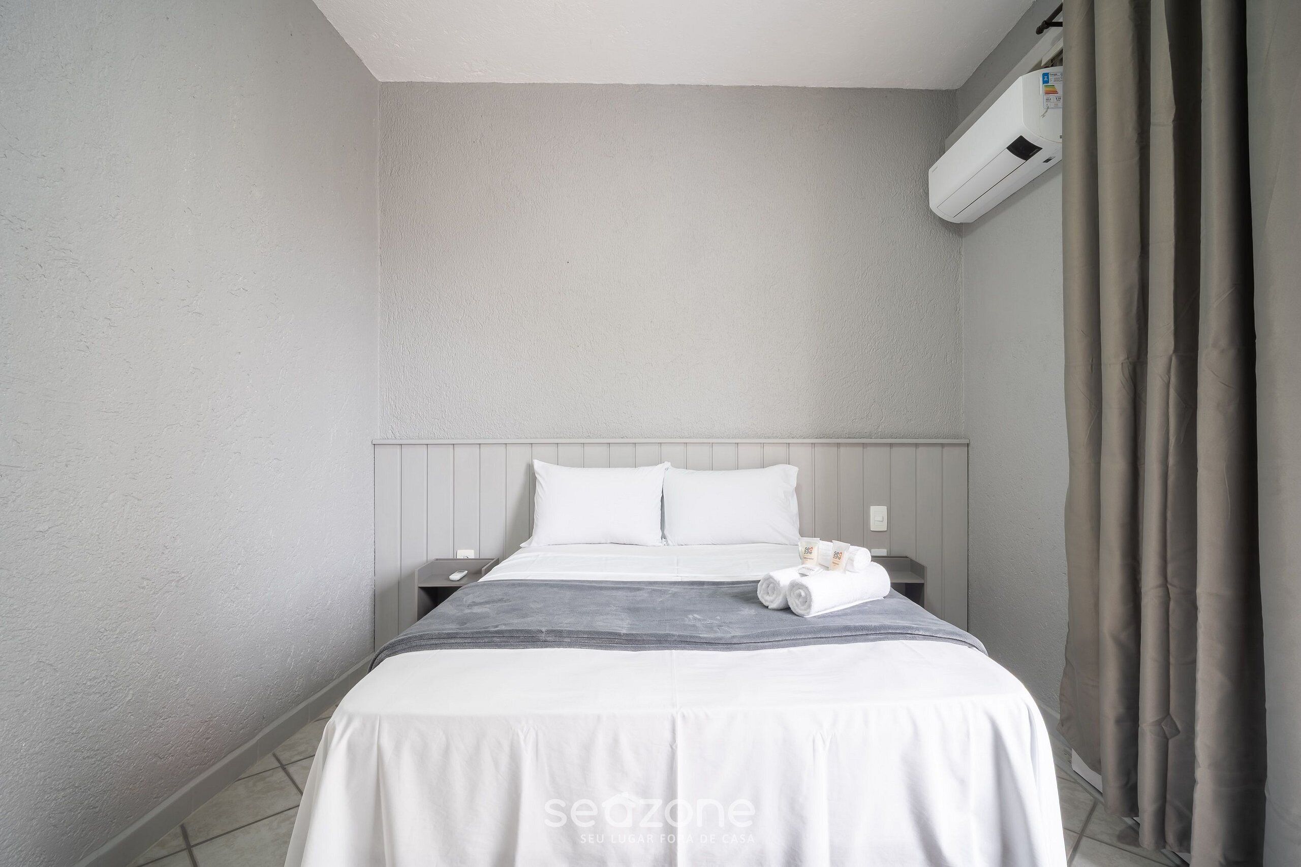 Una habitación de hotel con cama, mesita de noche, aire acondicionado instalado en la pared y cortina.