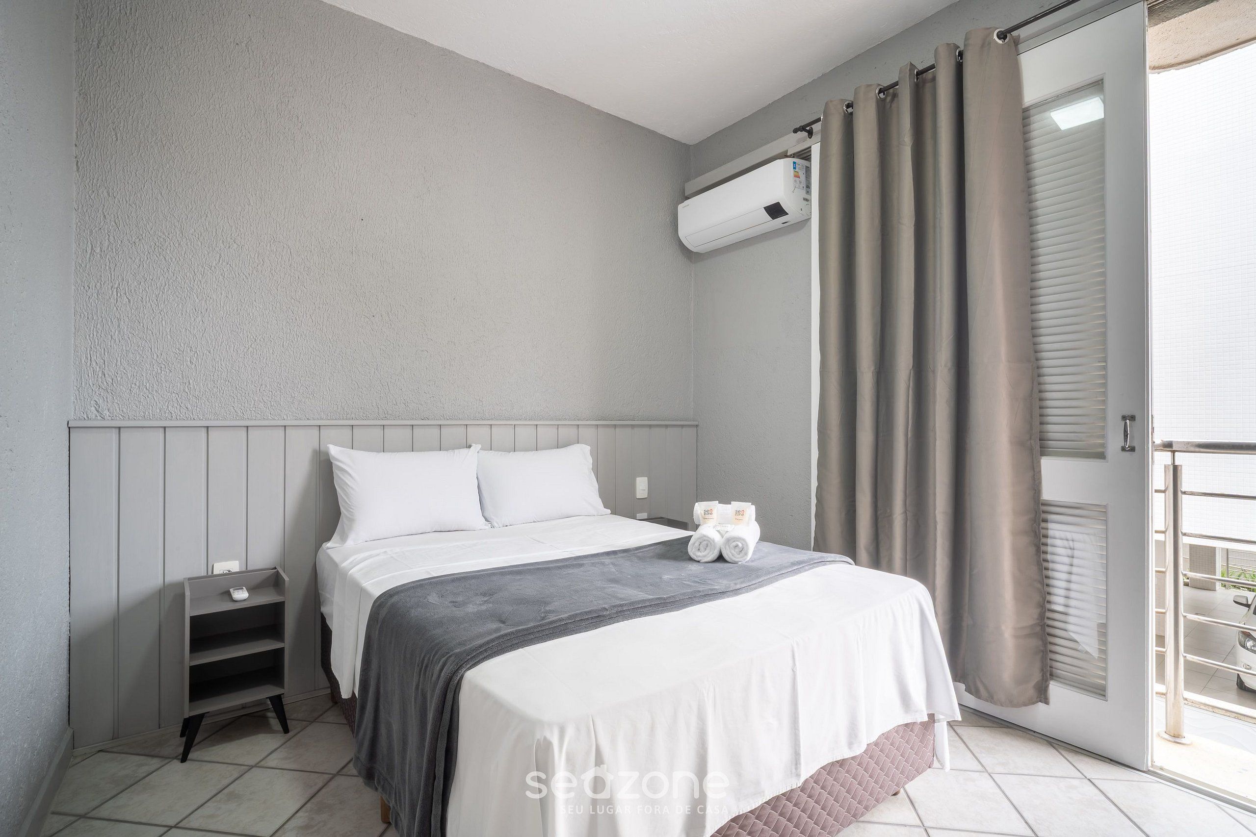A bedroom with a bed, a nightstand, an air conditioning unit, and a view of the outside.