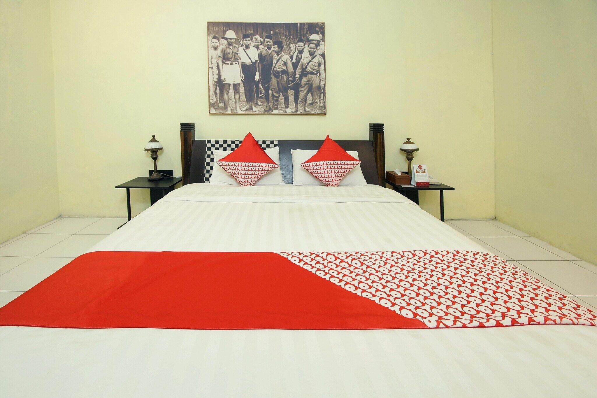 Habitación de hotel con una cama, dos mesitas de noche, una lámpara, un cuadro en la pared y un plaid rojo y blanco sobre la cama.