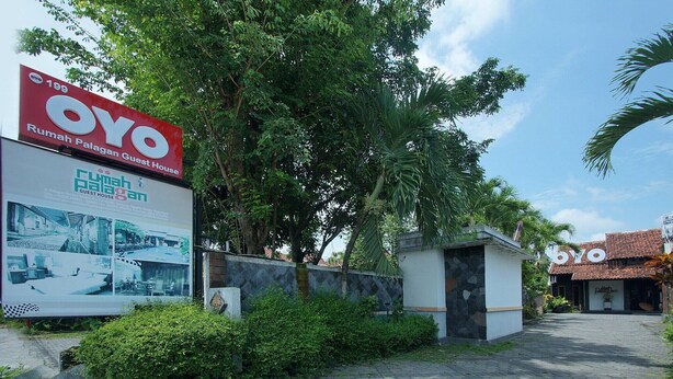 Een pad dat leidt naar een gebouw met een bordje voor 'OYO Guest House' en een grote reclameposter voor 'Rumah Palagen Guest House'.
