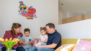 Een gezin zit op een gele bank, erboven een decoratie aan de muur.