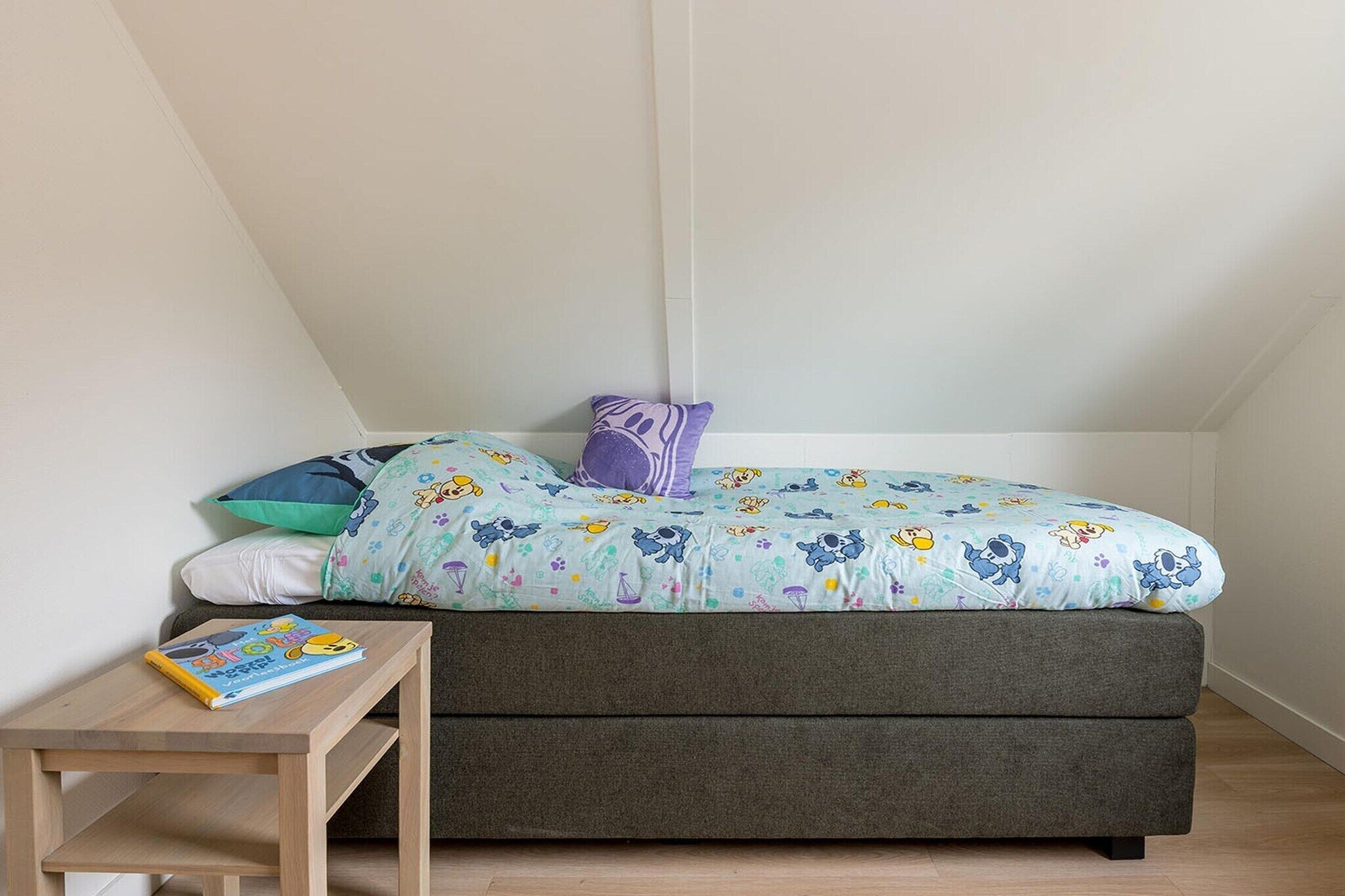 Um quarto pequeno e aconchegante com uma cama, travesseiros, uma mesinha de cabeceira e um livro.