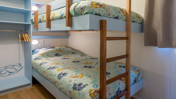 A bunk bed with a ladder, a shelf with hanging clothes, and a wall with a light switch.