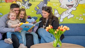 Eine Familie sitzt auf einer Couch, neben ihr ein Buch und ein Blumenstrauß mit Tulpen. Im Hintergrund ist ein Wandgemälde mit Cartoon-Motiven zu sehen.