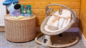 Una mecedora para bebés con asiento acolchado y panel de control, una cesta de mimbre con libros infantiles, y un peluche azul sobre un piso de madera.