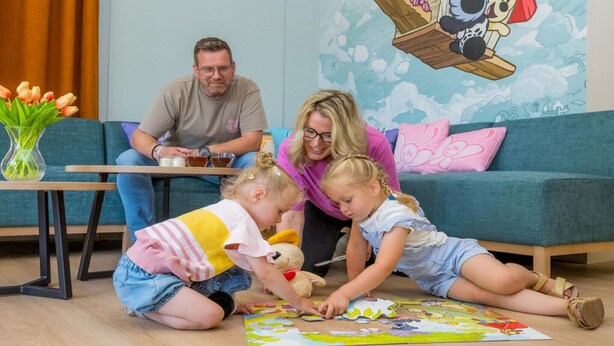 Eine Familie mit zwei Kindern sitzt auf dem Boden und spielt im Wohnzimmer mit einem Puzzle. Das Zimmer hat ein blaues Sofa und ein Wandbild.