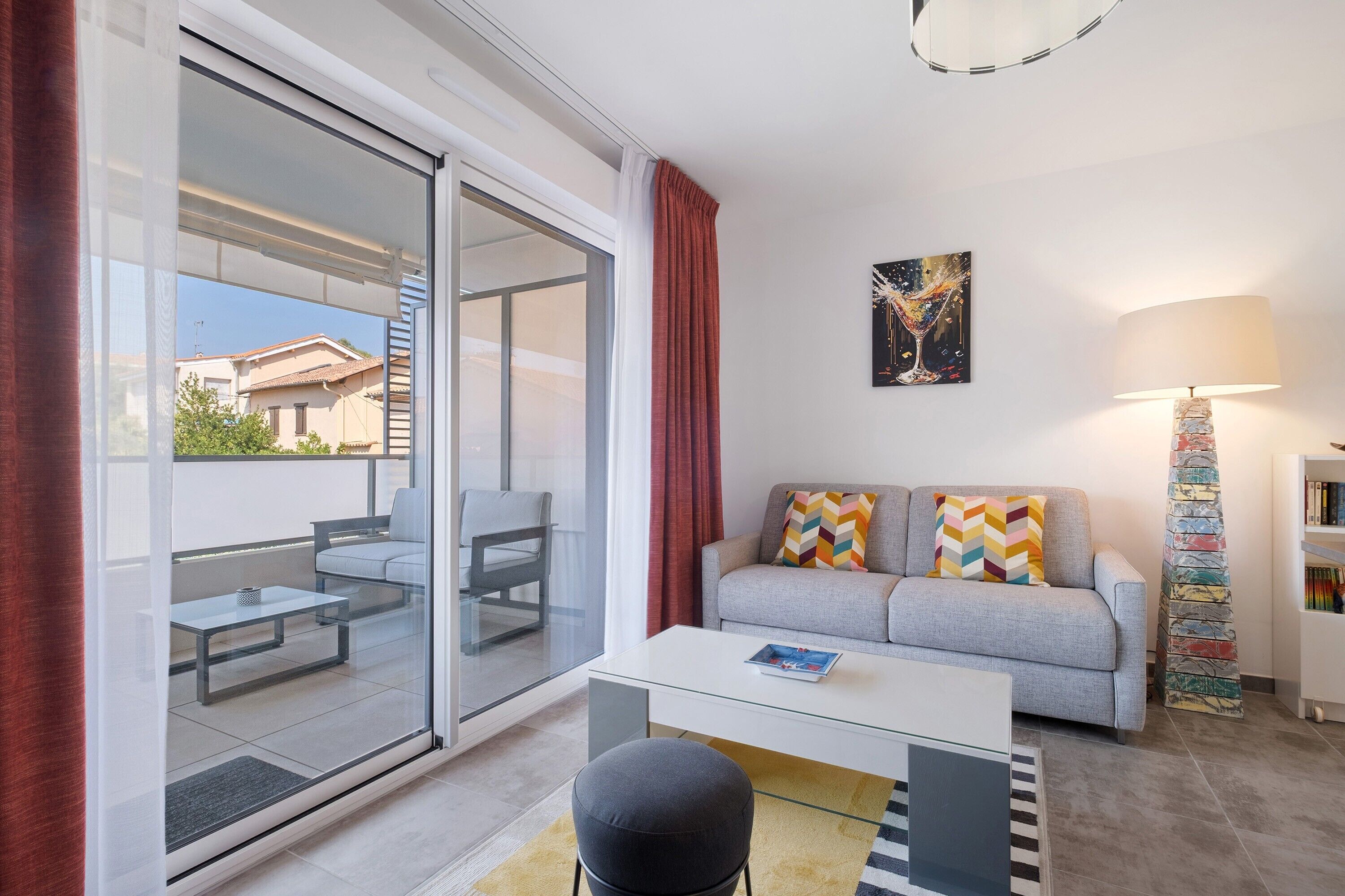 A modern living room with a glass door leading to a balcony, a grey sofa with colorful cushions, a white coffee table, and a lamp with a multicolored base.