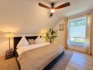 Ein Schlafzimmer mit einem Bett, einem Deckenventilator, einem Nachttisch, einer Lampe, einer Pflanze und einem Fenster mit Vorhängen.