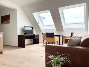 Ein modernes Wohnzimmer mit einer Couch, einem Schreibtisch mit Laptop und einem Fernseher. Im Deckenbereich befinden sich Dachfenster.