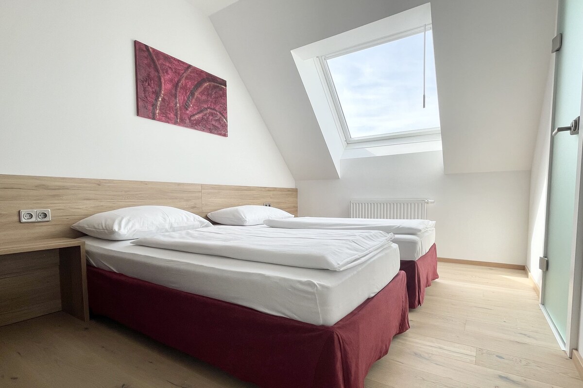 Un dormitorio con techo inclinado, tragaluces, cama con ropa de cama blanca y falda roja, y una obra de arte colgada en la pared.