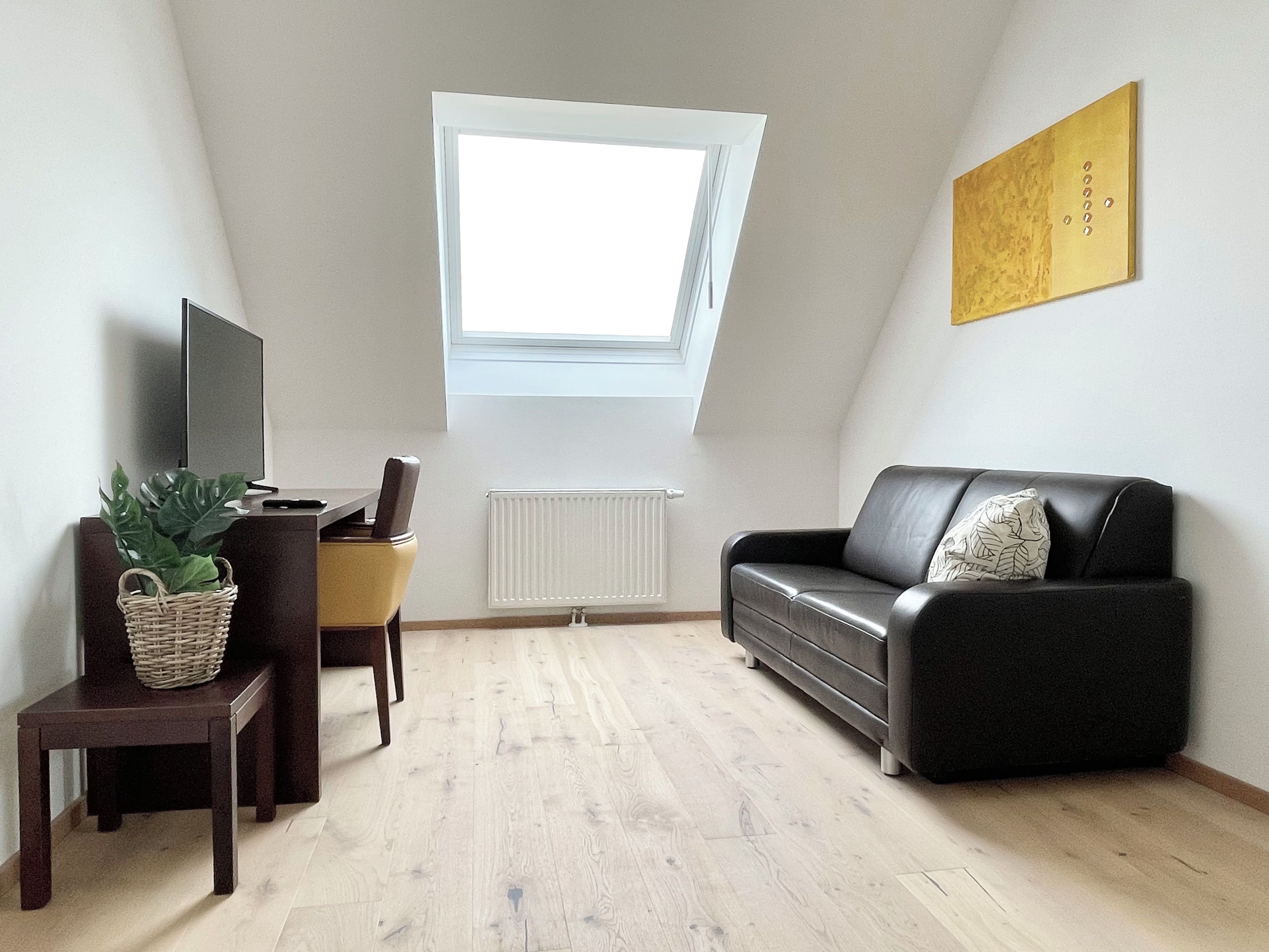 A room with a sloped ceiling, a black leather sofa, a television on a stand, a small table with a plant, and a radiator.