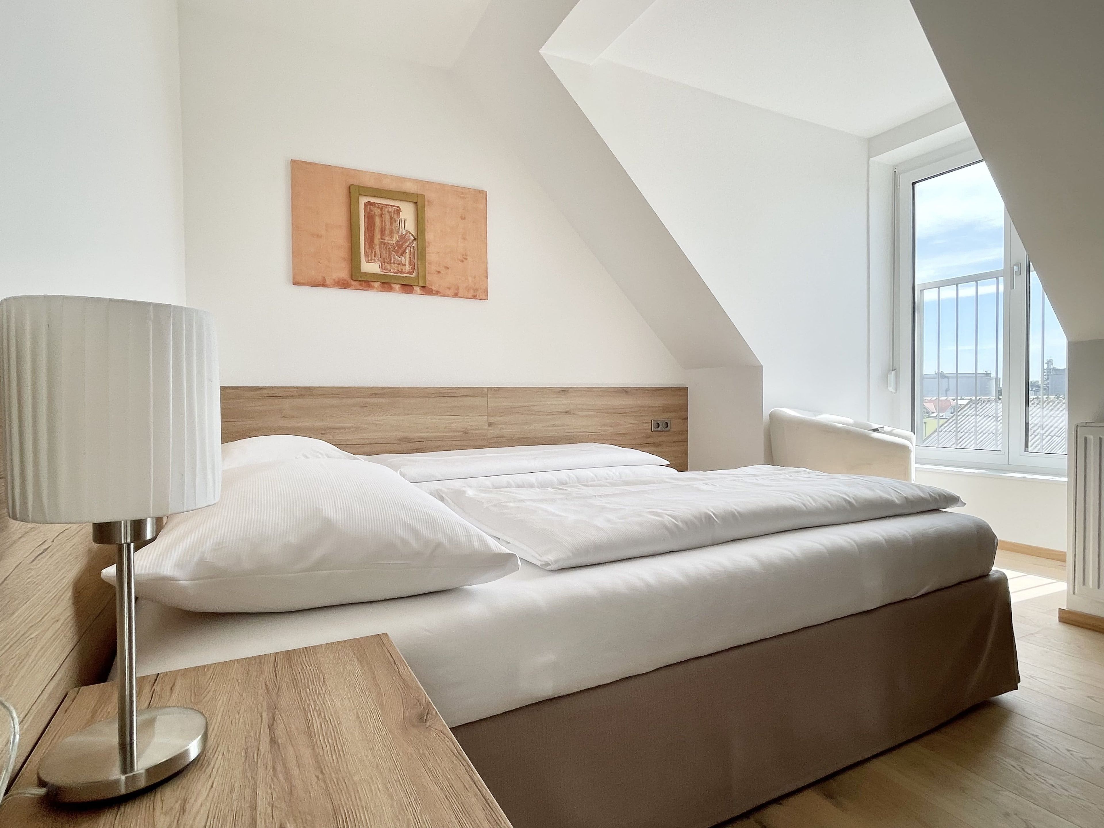A modern bedroom with a bed, a bedside lamp, a wooden headboard, a framed picture, and a large window.