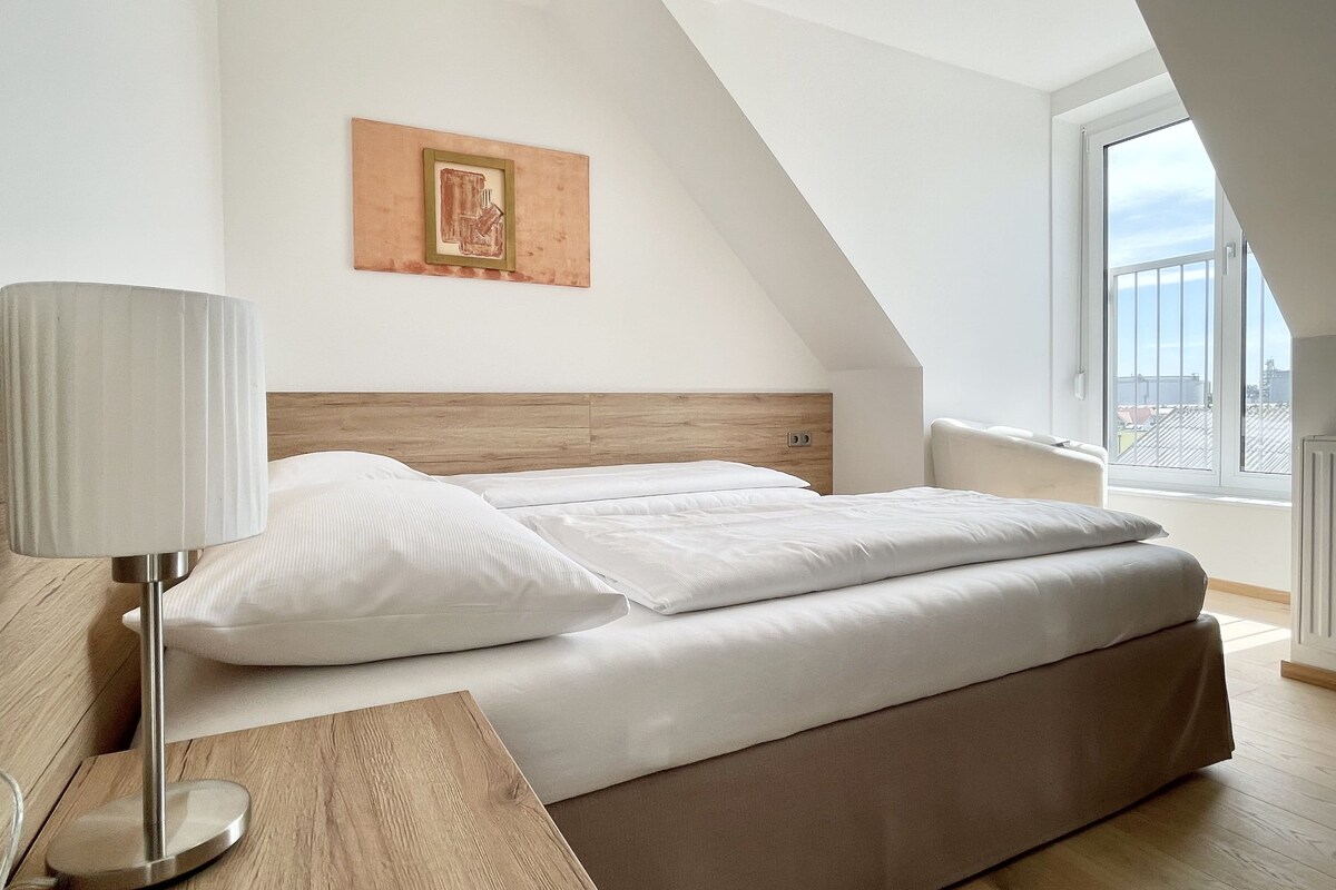Un dormitorio moderno con cama, lámpara de noche, cabecero de madera, un cuadro enmarco y un ventanal grande.