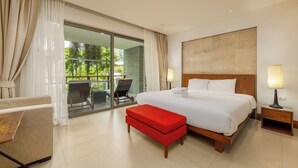Habitación de hotel con una cama grande, un banco rojo, un escritorio, una silla y un balcón con vistas a la vegetación.