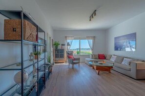 A modern living room with a sofa, coffee table, and shelves. There is a window with curtains and a view of the outdoors.