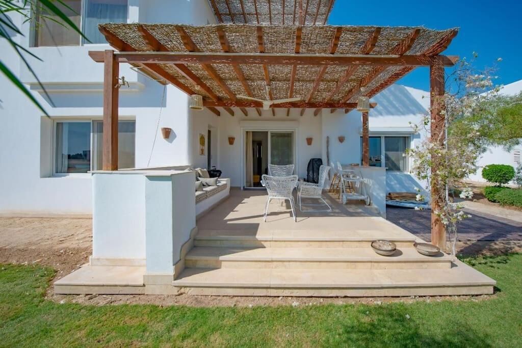 A covered outdoor seating area with a pergola, steps leading to a door, and a white building in the background.